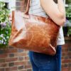 A person wearing a striped top and blue jeans stands outdoors near flowers and a brick wall, carrying the Personalised Brown Leather Zipped Tote Handbag over their shoulder.