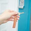 A hand holds a key with a Personalised Brown Leather Tag Keyring labeled "OUR HOME" in front of a door with a silver lock and blue trim.