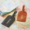 Two Personalised Leather Luggage Tags—one black labelled "T. SMITH," the other brown with "G.R.V"—sit on a map beside an open passport, ready for stylish travel with your own custom touch.