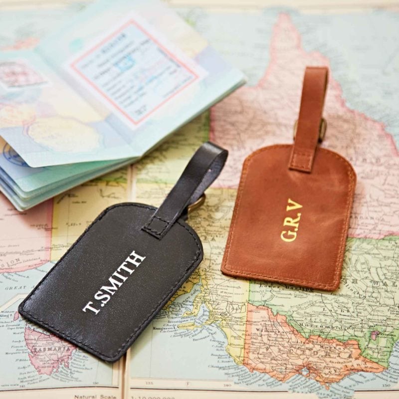 Two Personalised Leather Luggage Tags—one black labelled "T. SMITH," the other brown with "G.R.V"—sit on a map beside an open passport, ready for stylish travel with your own custom touch.