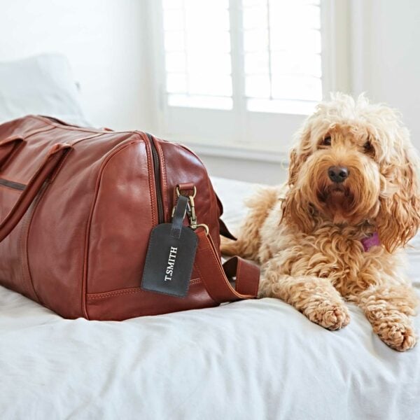 A Personalised Leather Luggage Tag reading "T. SMITH" is attached to a brown leather duffel bag, which rests on a bed beside a fluffy, light-colored dog.