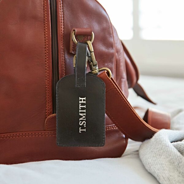 A brown leather bag with a personalised leather luggage tag attached, displaying the name "T. SMITH" in white capital letters.