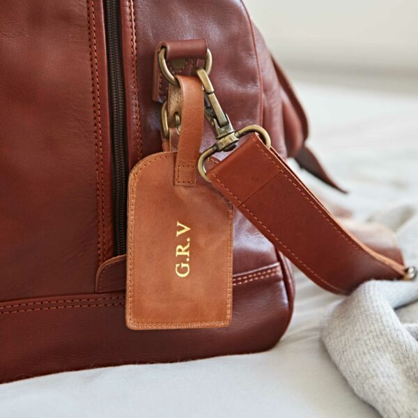 Close-up of a brown leather bag with a personalised leather luggage tag attached, displaying the initials "G.R.V." in gold letters.