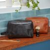 Two Personalised Leather Wash Bags—one black monogrammed "C.H.R" and one brown with "HASAN"—rest on a bathroom counter beside a toothbrush and shaving brush.