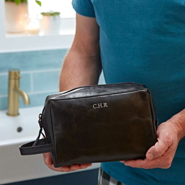 A person in a blue shirt holds a personalised leather wash bag, with the initials "C.H.R" embossed on the front, standing in a bathroom.
