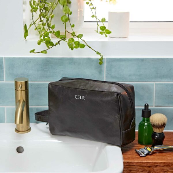 A Personalised Leather Wash Bag with “C.H.R” initials rests on a bathroom counter beside a sink, shaving brush, razor, green bottle, and plant.
