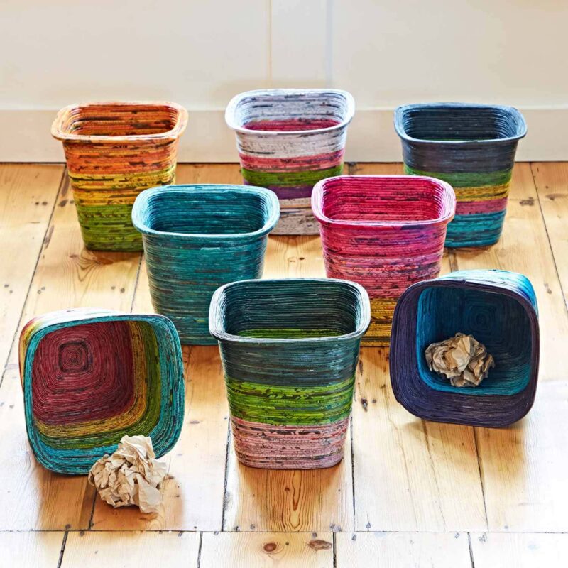 Seven vibrant Recycled Newspaper Waste Paper Baskets are arranged on a wooden floor, accompanied by two crumpled paper balls.
