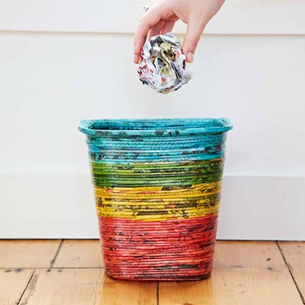 A hand is throwing a crumpled piece of recycled newspaper into a colorful, multi-striped Recycled Newspaper Waste Paper Basket on a wooden floor.