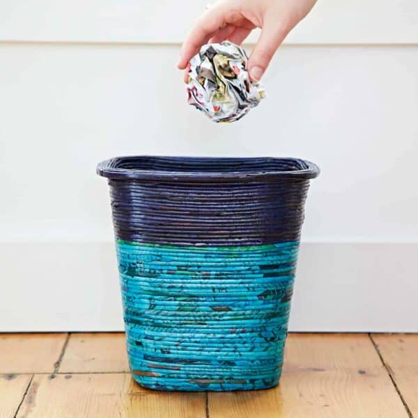 A hand is dropping a crumpled piece of newspaper into a blue and black Recycled Newspaper Waste Paper Basket on a wooden floor.