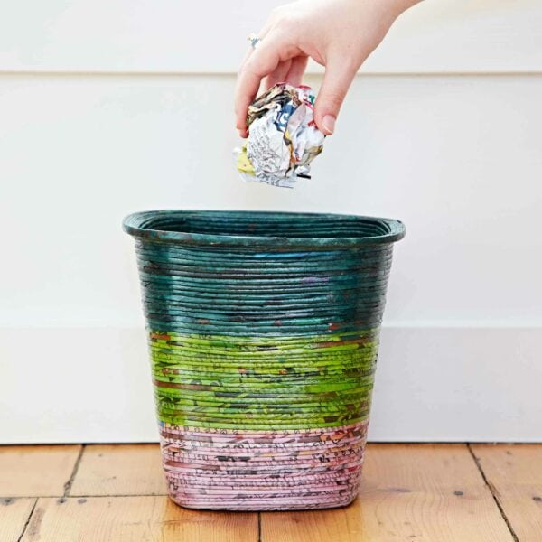 A hand drops a crumpled piece of recycled newspaper into a Recycled Newspaper Waste Paper Basket placed on a wooden floor.
