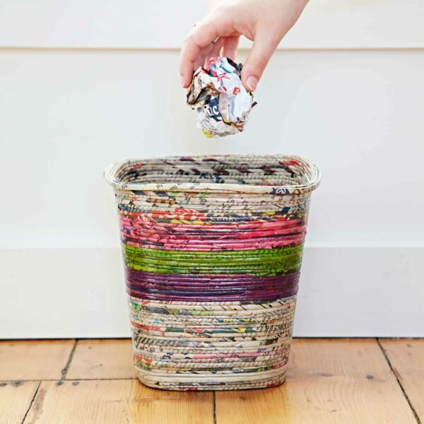 A hand is placing a crumpled piece of colorful recycled paper into a Recycled Newspaper Waste Paper Basket set on a wooden floor against a plain wall.