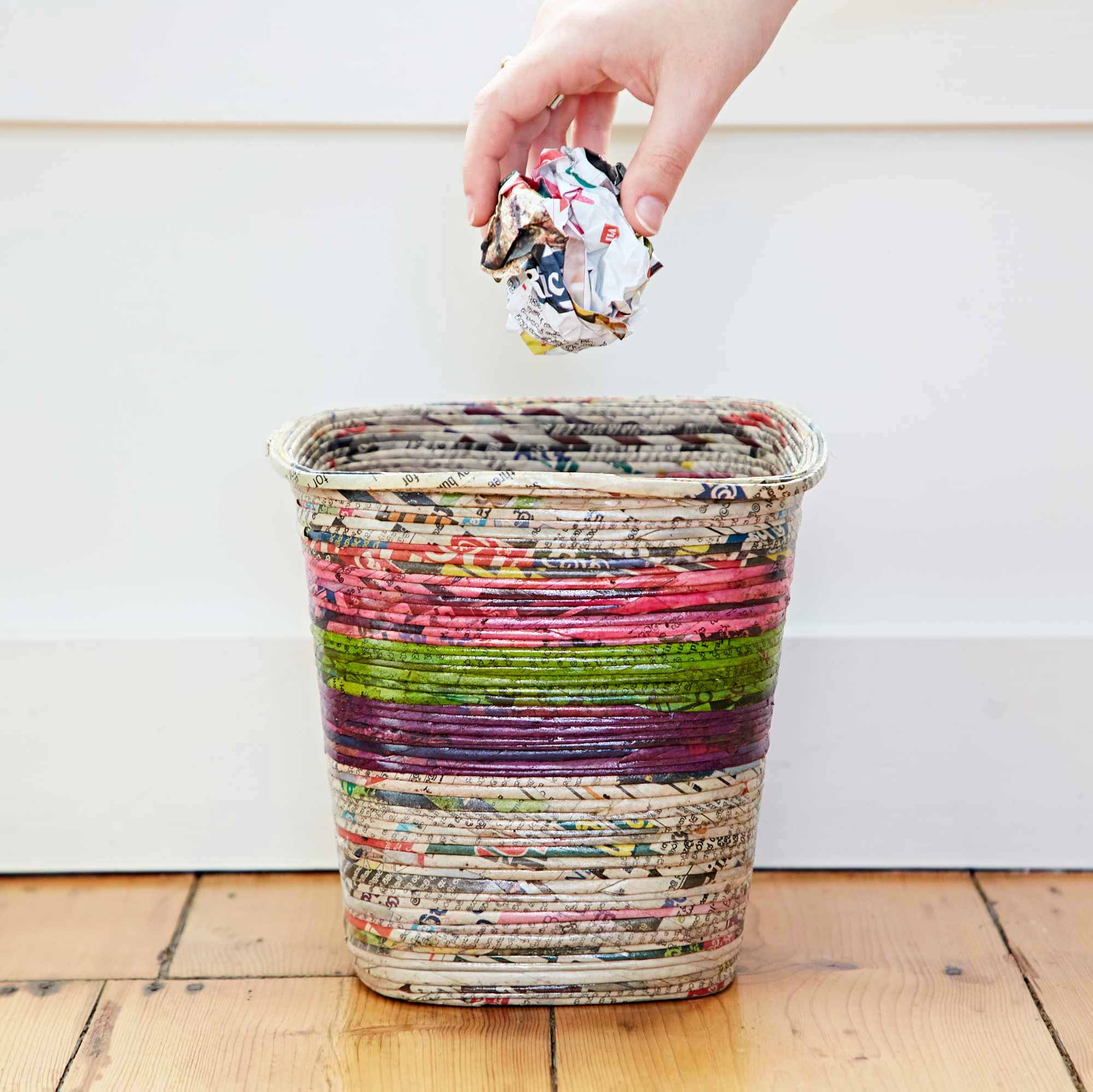 A hand is placing a crumpled piece of colorful recycled paper into a Recycled Newspaper Waste Paper Basket set on a wooden floor against a plain wall.