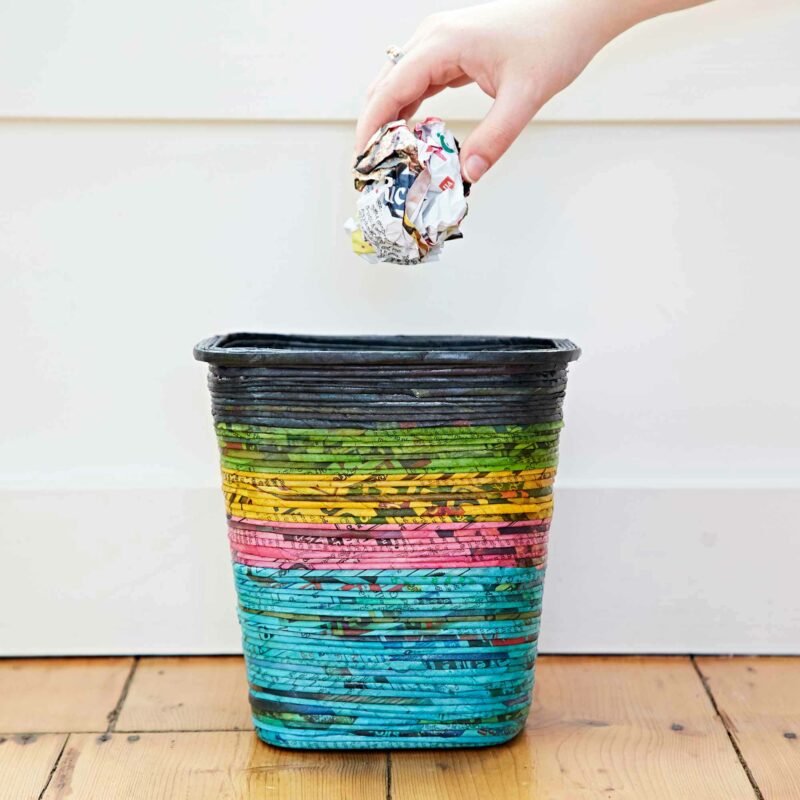 A hand dropping a crumpled piece of paper into a Recycled Newspaper Waste Paper Basket on a wooden floor.