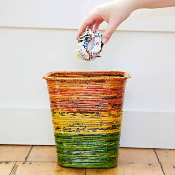 A hand drops a crumpled piece of paper into a Recycled Newspaper Waste Paper Basket.