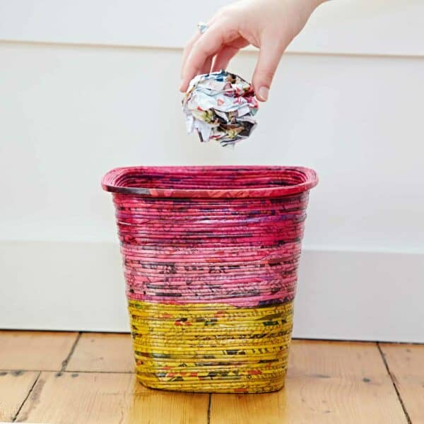 A hand drops a crumpled paper ball into a Recycled Newspaper Waste Paper Basket with pink and yellow hues, placed on a wooden floor.