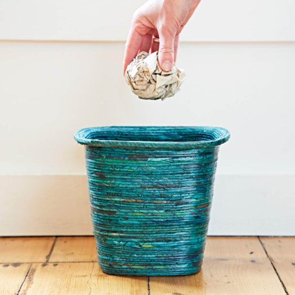 A hand discards crumpled newspaper into a Recycled Newspaper Waste Paper Basket on a wooden floor.