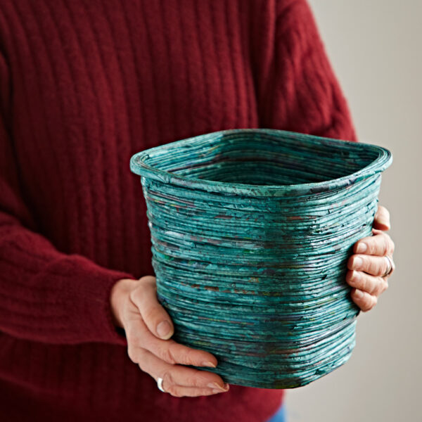 A person wearing a red jumper holds a square, teal Recycled Newspaper Waste Paper Bin made from layered recycled newspaper.