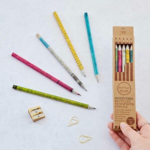 A hand holds a box of Recycled Newspaper Pencils. Five pencils are displayed in various colors alongside a pencil sharpener and two paper clips on a light surface.