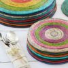 Stacked colorful woven coasters with a set of silver utensils wrapped in a beige napkin next to a stack of Round Recycled Newspaper Placemats on a white wooden table.