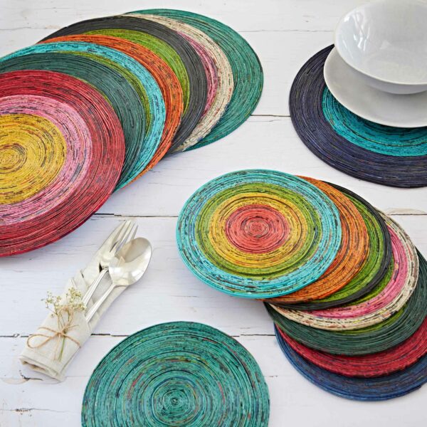 A variety of colorful, Round Recycled Newspaper Placemats are arranged on a white wooden table. Nearby are white plates stacked on a dark placemat and wrapped silverware with a small flower.
