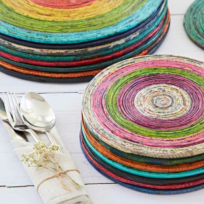 Stacked colorful woven coasters with a set of silver utensils wrapped in a beige napkin next to a stack of Round Recycled Newspaper Placemats on a white wooden table.