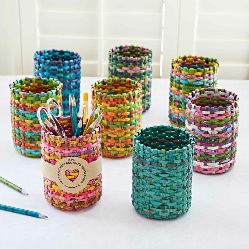 A collection of colorful Round Recycled Newspaper Pencil Holders sits on a white table, each one made from 100% recycled newspaper and holding various writing utensils.