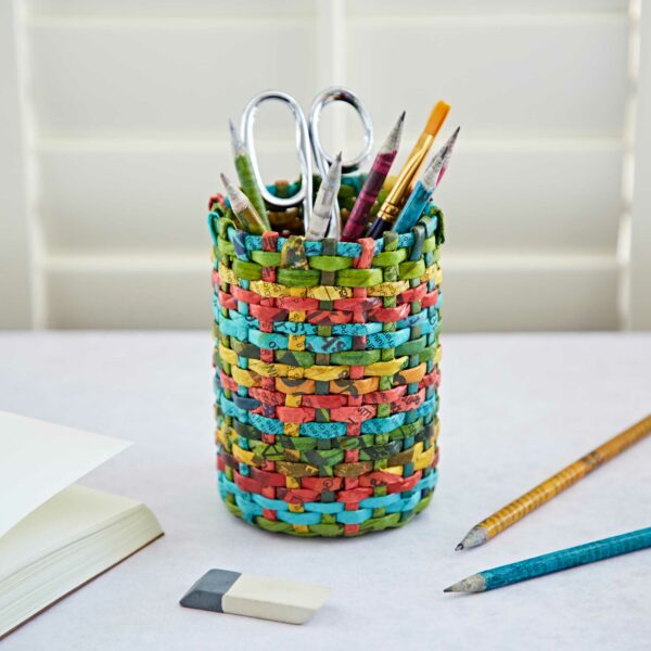 A colorful Round Recycled Newspaper Pencil Holder on a desk contains scissors, pencils, and brushes, with additional pencils, an eraser, and an open notebook nearby.