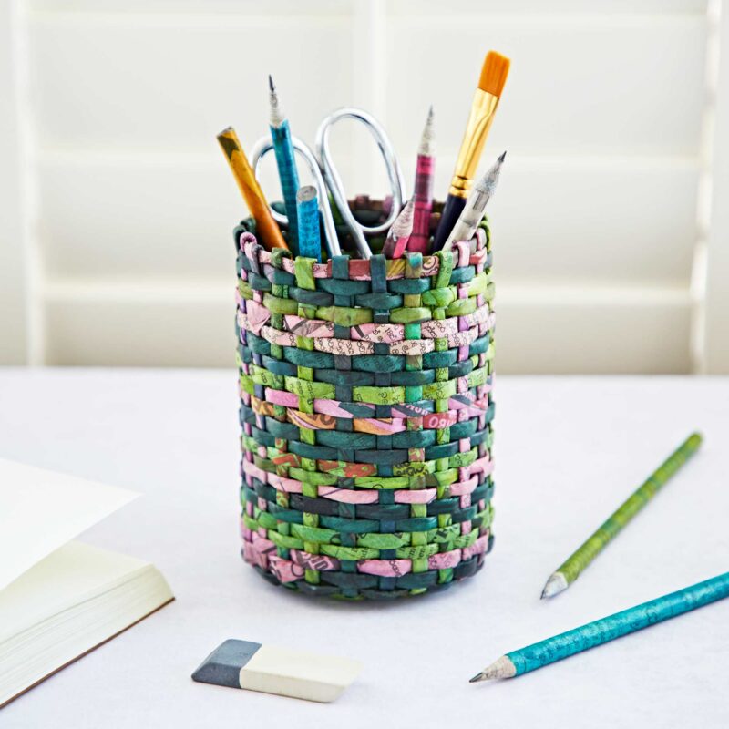 A Round Recycled Newspaper Pencil Holder holds various art supplies—including brushes, scissors, and pencils—sitting on a white desk beside a book, eraser, and two more pencils.