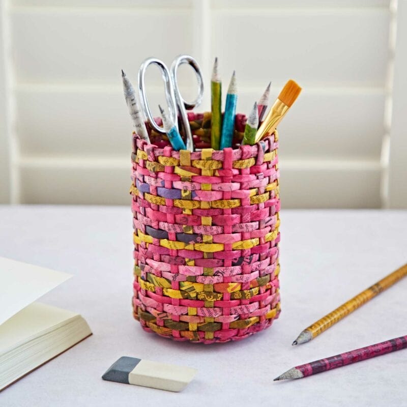 A Round Recycled Newspaper Pencil Holder, made from recycled newspaper, contains various stationery items, including pencils, pens, scissors, and a paintbrush. It is placed on a white surface next to an eraser and a notebook.