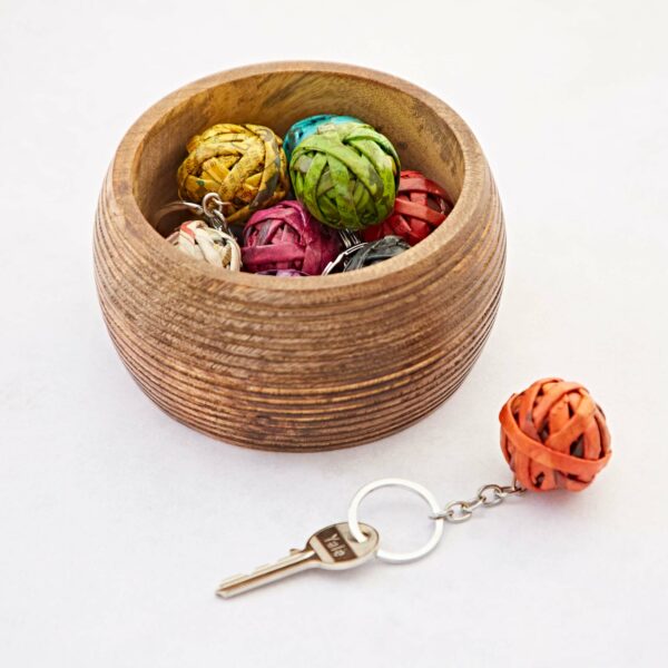 A wooden bowl contains several colorful woven balls, one of which is attached to a Recycled Newspaper Ball Keyring, placed outside the bowl. The key has a metal tag with the number 740, adding a touch of intrigue.