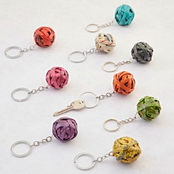 A collection of colorful Recycled Newspaper Ball Keyrings made from woven, recycled material is displayed on a flat surface. A single key is attached to one of the keyrings.