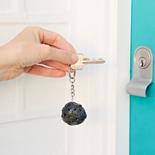 A hand is holding a key with a Recycled Newspaper Ball Keyring, inserting it into a lock on a teal-colored door.