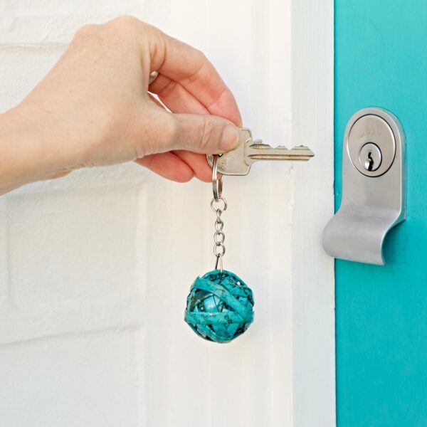A hand is inserting a key with a Recycled Newspaper Ball Keyring into the keyhole of a turquoise door.