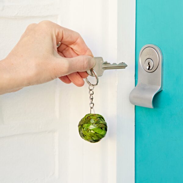 A hand is inserting a key with a Recycled Newspaper Ball Keyring into a cylindrical door lock on a turquoise door.