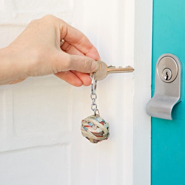 Hand holding a key with a Recycled Newspaper Ball Keyring, inserting the key into a lock on a turquoise door. The keyring looks sturdy and stylish, made from recycled materials.