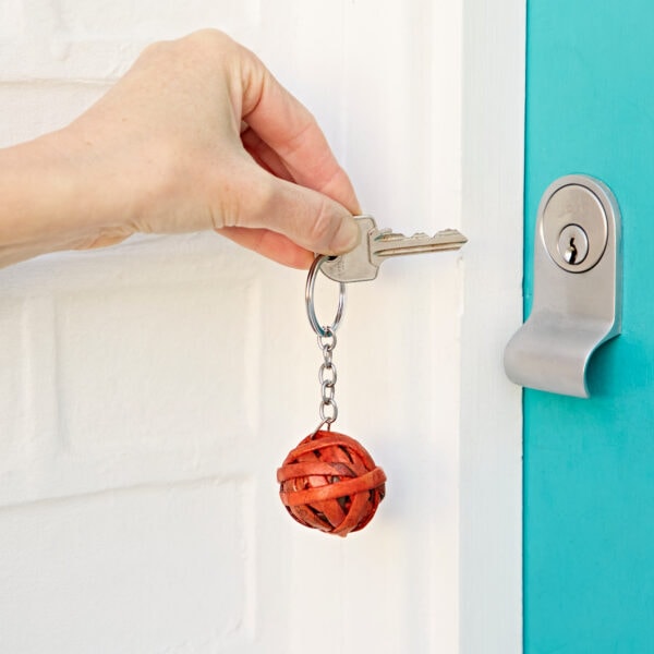 A hand inserts a key into a teal door lock. The keychain, featuring a decorative Recycled Newspaper Ball Keyring, adds a touch of personality to the scene.