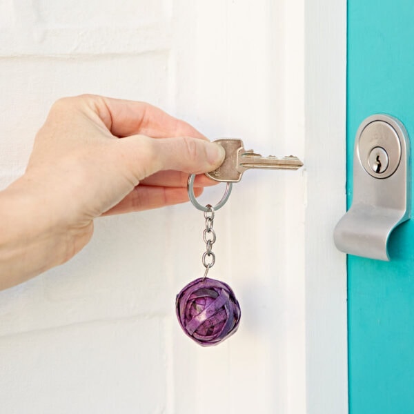 A hand is holding a key with a Recycled Newspaper Ball Keyring, inserting it into a door lock framed by a turquoise door.
