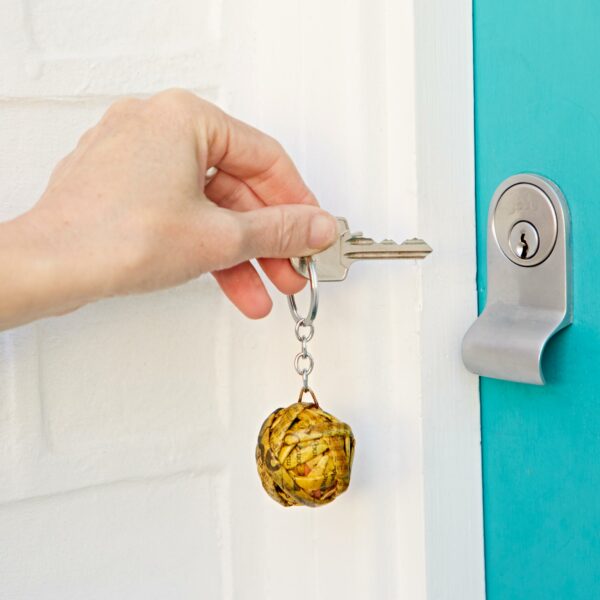 A hand holding a key with an artichoke keychain inserts it into a turquoise door lock, the Recycled Newspaper Ball Keyring adding a touch of eco-chic.