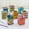 Colorful, recycled newspaper desk organizers of various sizes and patterns displayed on a white surface, accompanied by pencils and a Square Recycled Newspaper Pencil Holder filled with stationery items.