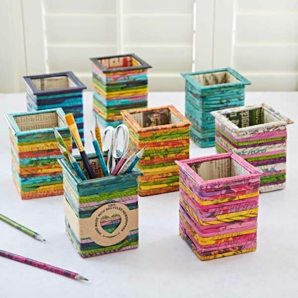 Colorful, recycled newspaper desk organizers of various sizes and patterns displayed on a white surface, accompanied by pencils and a Square Recycled Newspaper Pencil Holder filled with stationery items.
