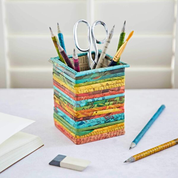 A Square Recycled Newspaper Pencil Holder containing pencils and scissors sits on a desk alongside an open notebook and erasers.