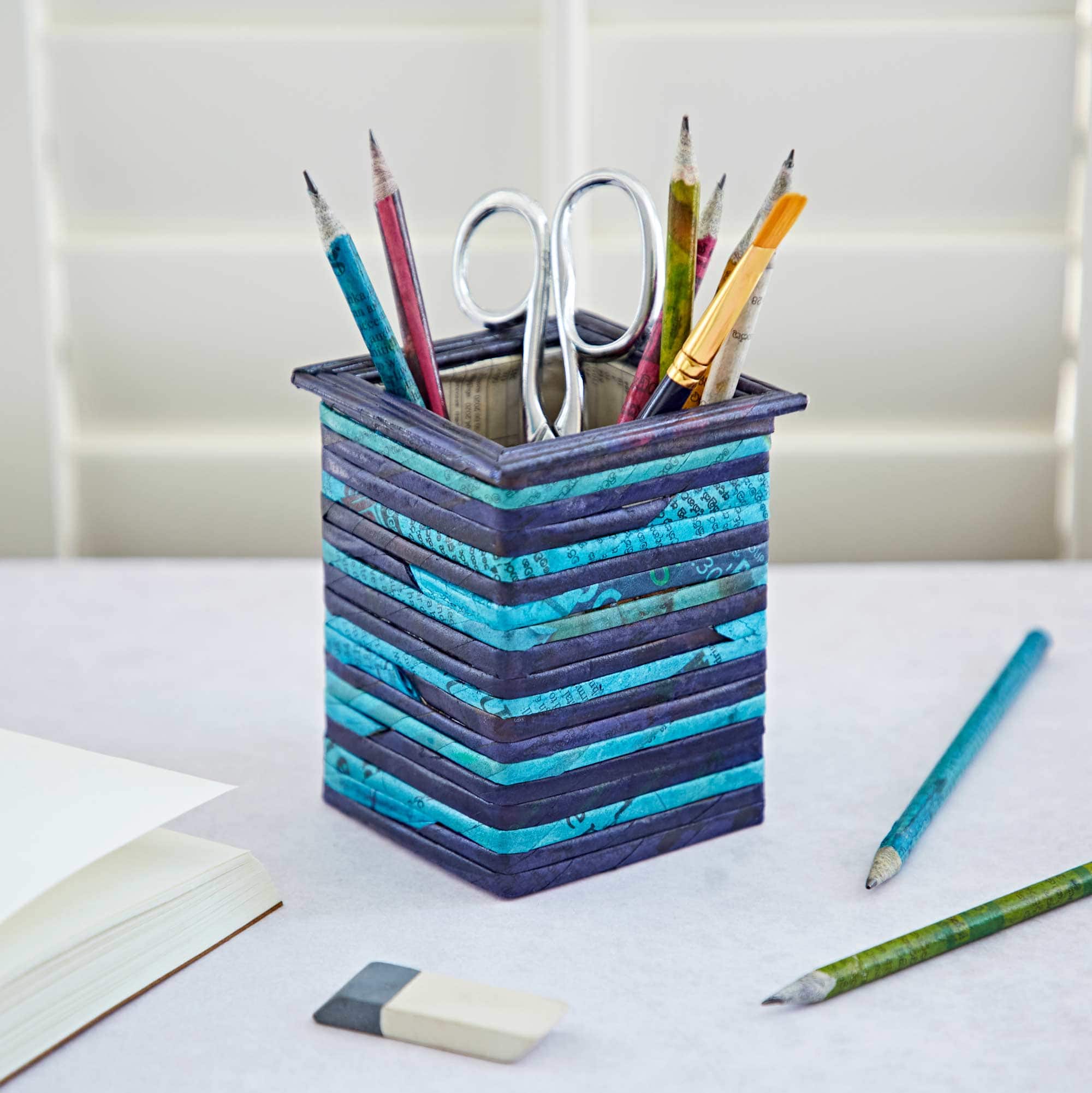 A Square Recycled Newspaper Pencil Holder contains pencils and scissors. It sits on a white desk next to an open book, two pencils, and an eraser.