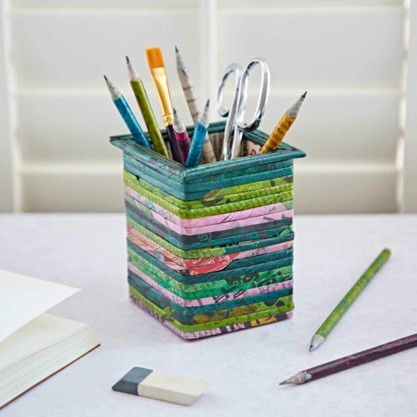 A colorful, striped Square Recycled Newspaper Pencil Holder sits on a desk, holding pencils, brushes, and scissors. An eraser, a green pencil, and an open notebook are placed nearby.