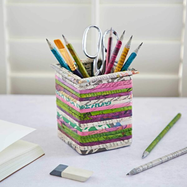A Square Recycled Newspaper Pencil Holder holds pencils, pens, scissors, and a ruler. An eraser, pencil, and open notebook are beside it on a white surface against a background of white blinds.