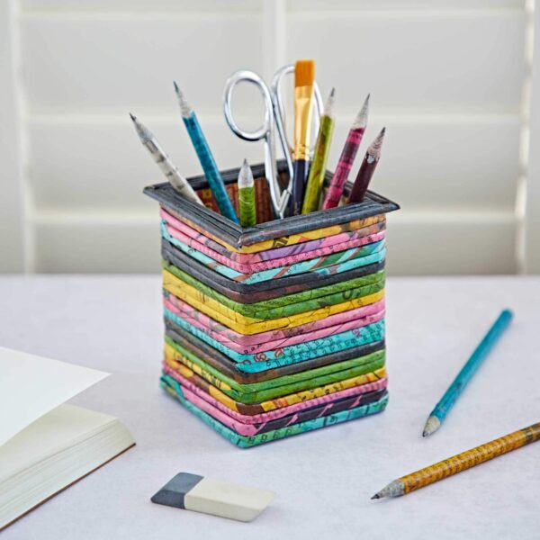 Square Recycled Newspaper Pencil Holder on a desk containing pencils, brushes, and scissors, next to a notebook, an eraser, and additional pencils. The square recycled newspaper design adds an eco-friendly touch. Background includes window blinds.