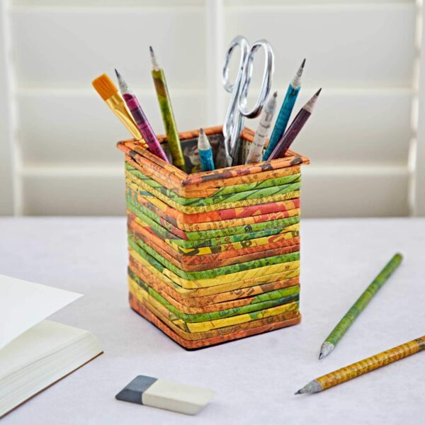 A Square Recycled Newspaper Pencil Holder containing scissors, pencils, pens, and a paintbrush sits on a white table next to an eraser, two pencils, and a partially open notebook.