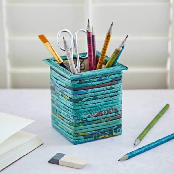 A Square Recycled Newspaper Pencil Holder holds pencils, a paintbrush, and scissors next to an eraser and an open notebook.