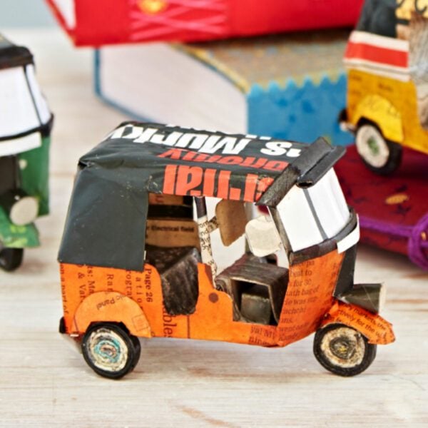 The Recycled Newspaper Tuk Tuk is a small, eco-friendly auto-rickshaw model crafted from recycled paper and metal, featuring orange and black colors, displayed on a light wooden surface.