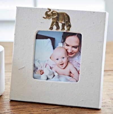 A photo frame featuring a mother and baby, topped with an elegant elephant ornament made from elephant dung, adorns a smooth wooden surface. Known as the Elephant Dung Photo Frame.