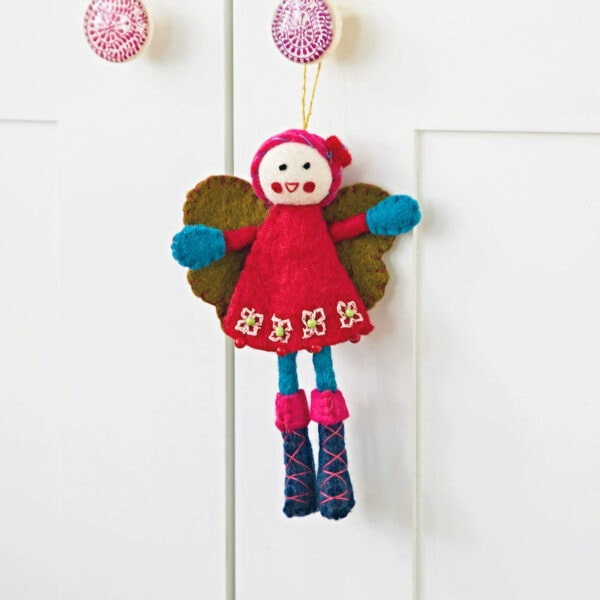 The Felt Alpine Girl Decoration, featuring a vibrant red dress, green wings, and blue boots, is hanging on a white cabinet.